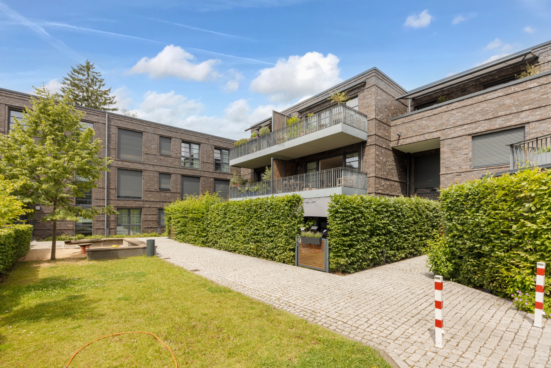 Moderner Wohnkomplex mit Balkonen, Garten und gepflastertem Weg, umgeben von Hecken.