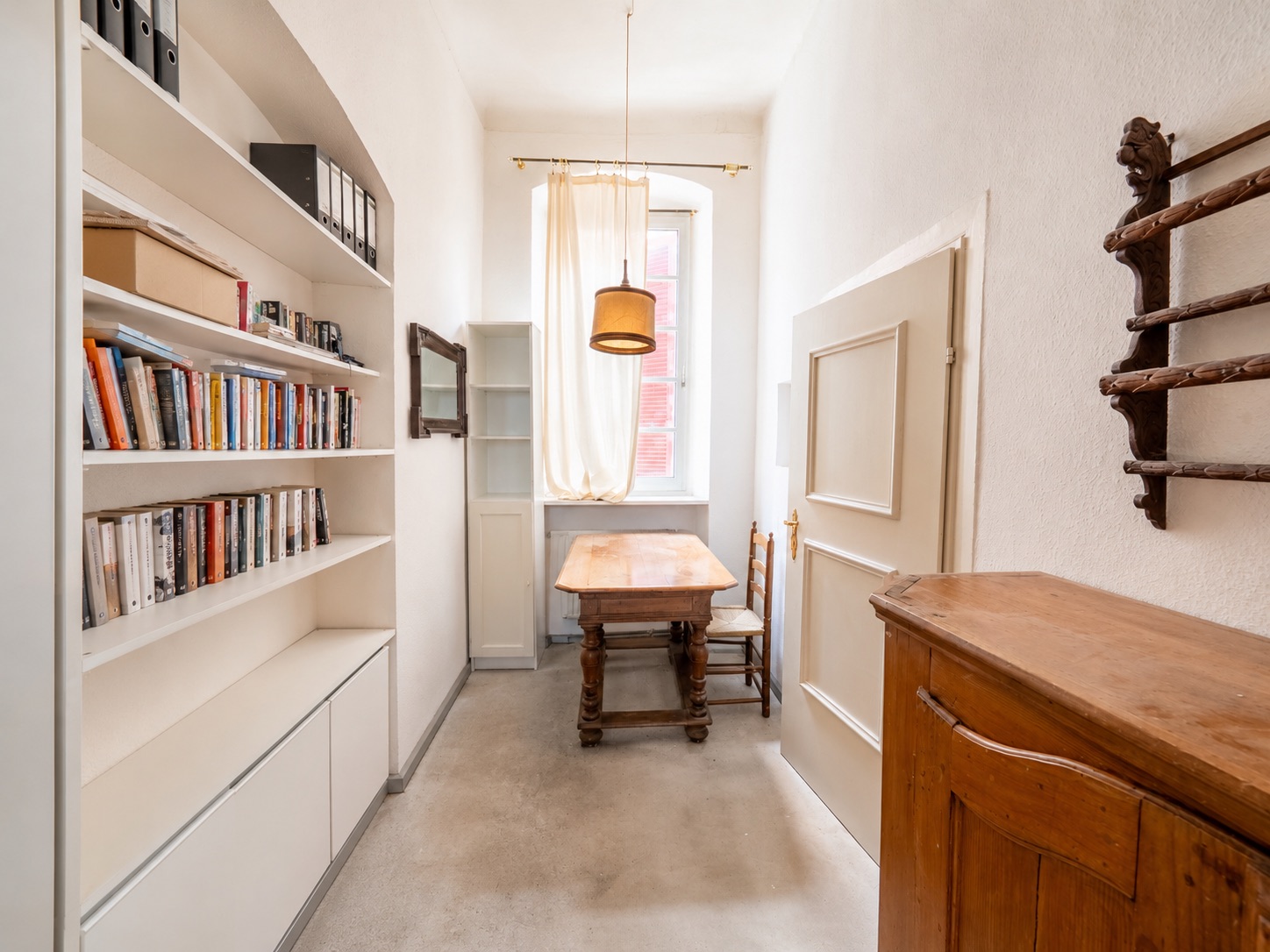 Heller Raum mit Bücherregal, Holztisch, zwei Stühlen und Fenster mit Vorhang.