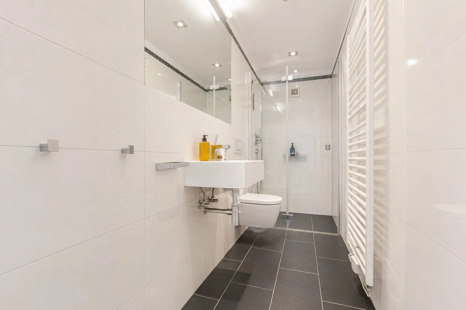 Modernes Badezimmer mit weißen Fliesen, Wandwaschbecken, WC, Handtuchheizkörper und Dusche.
