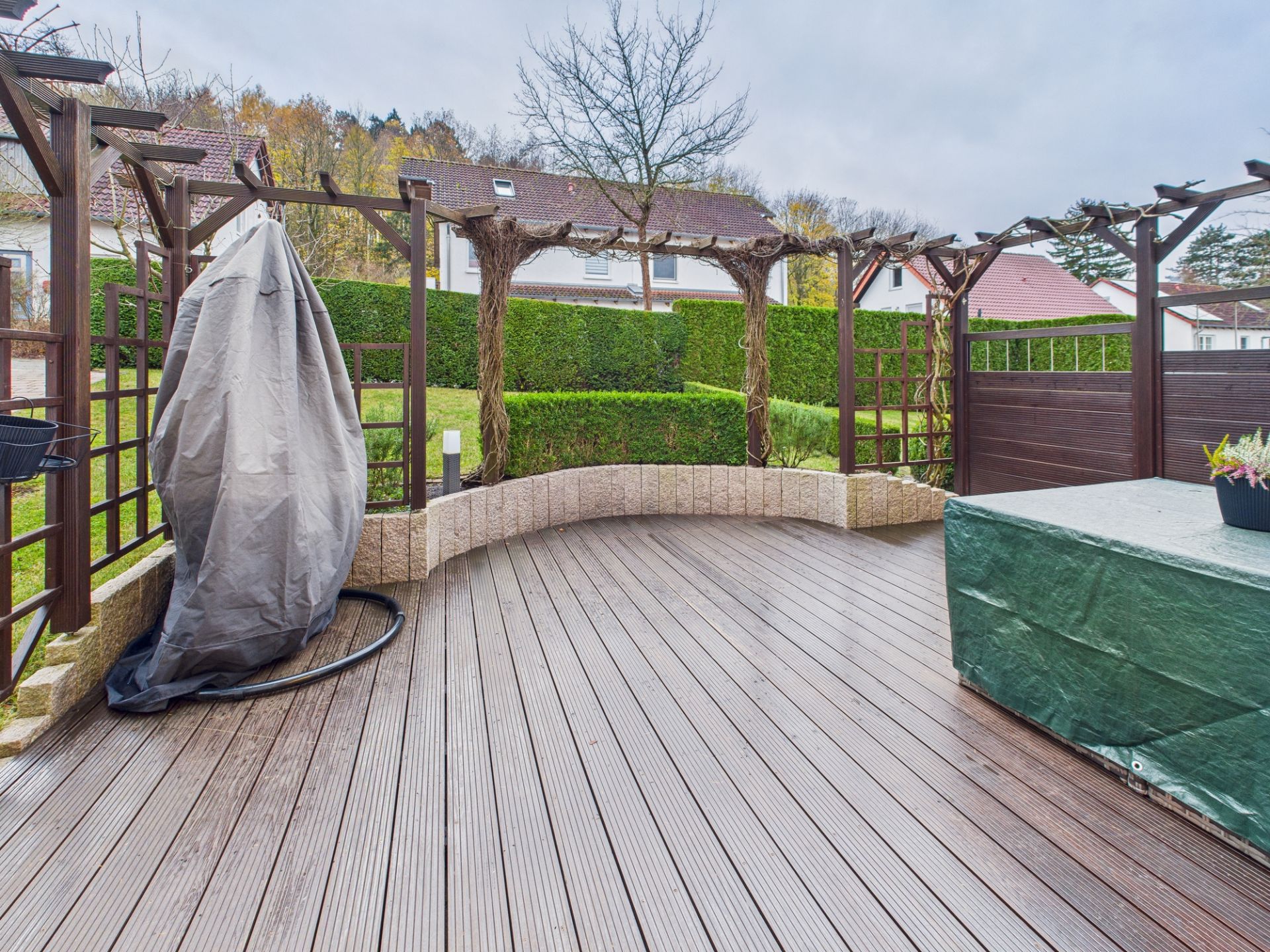 Überdachte Holzterrasse mit Rankgittern und abgedeckten Gartenmöbeln, Garten und Hecken im Hintergrund.