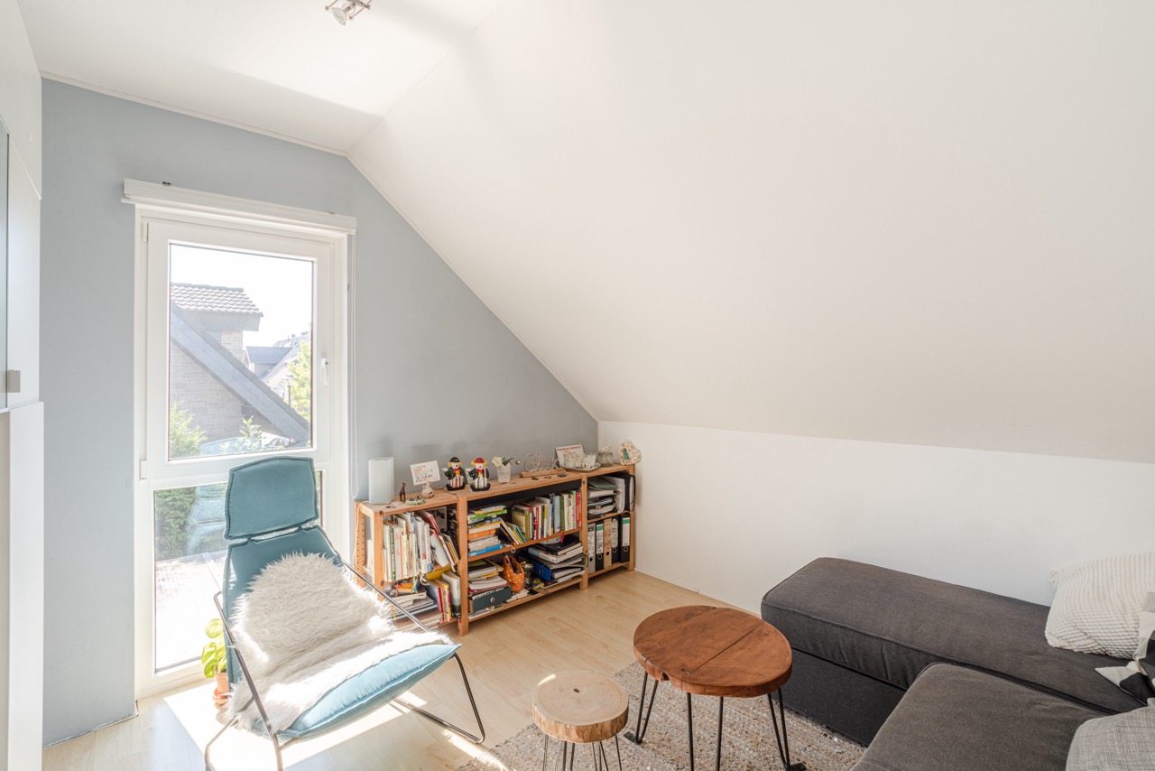 Helles Wohnzimmer mit schräger Decke, Sessel, Regal, Sofa und kleinen Tischen.