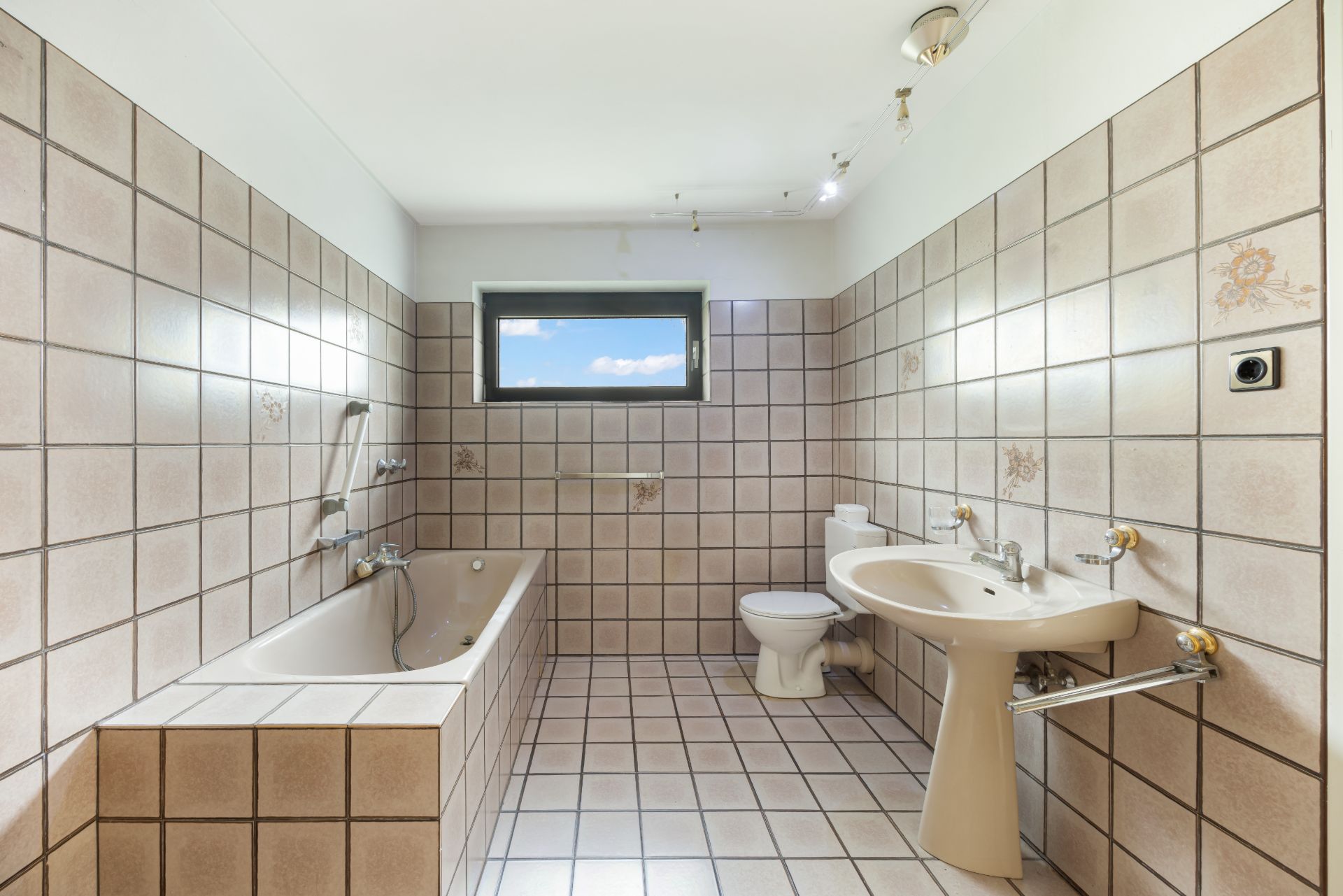 Geräumiges Badezimmer mit Badewanne, WC, Waschbecken und kleinem Fenster, gefliest in Beige.