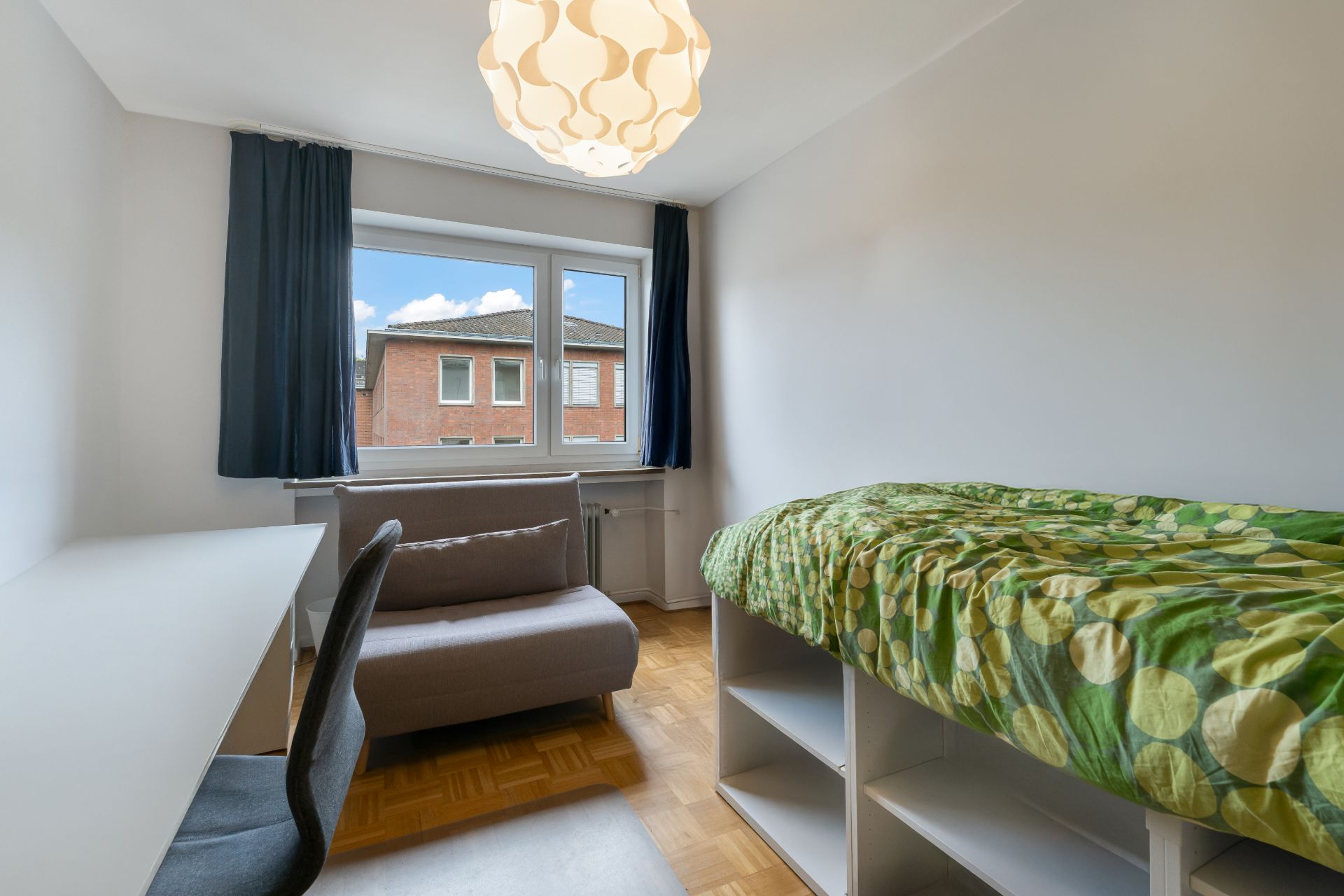 Helles Zimmer mit Schreibtisch, Stuhl, Sofa und Bett mit grüner Decke.