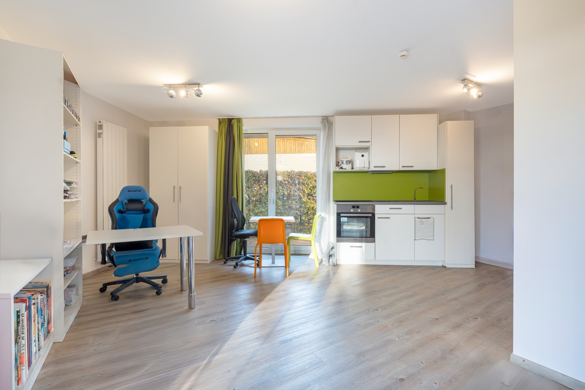 Helles Wohnstudio mit Schreibtisch, Regal, Küchenzeile und Essbereich mit bunten Stühlen.