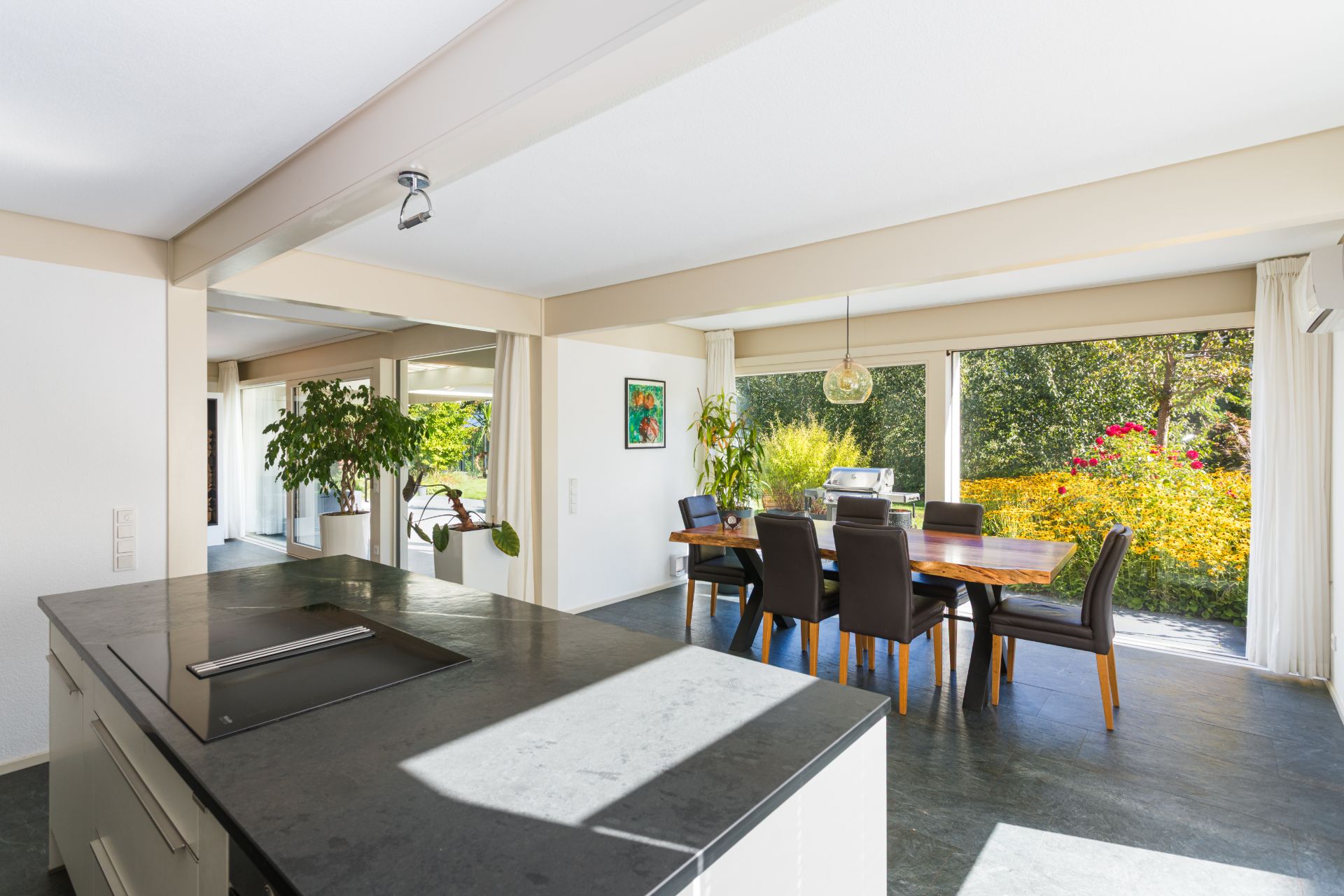 Moderne Küche mit Kochinsel und Essbereich mit großem Holztisch, hellem Boden und großen Fenstern zum Garten.