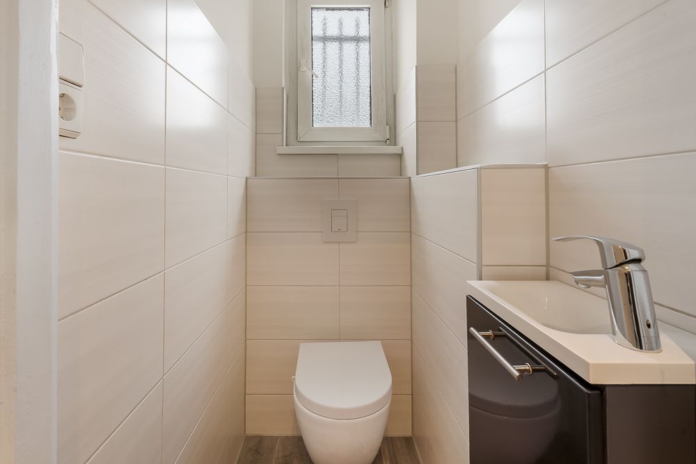 Schmaler, moderner WC-Raum mit Toilette, kleinem Waschbecken und Fenster.