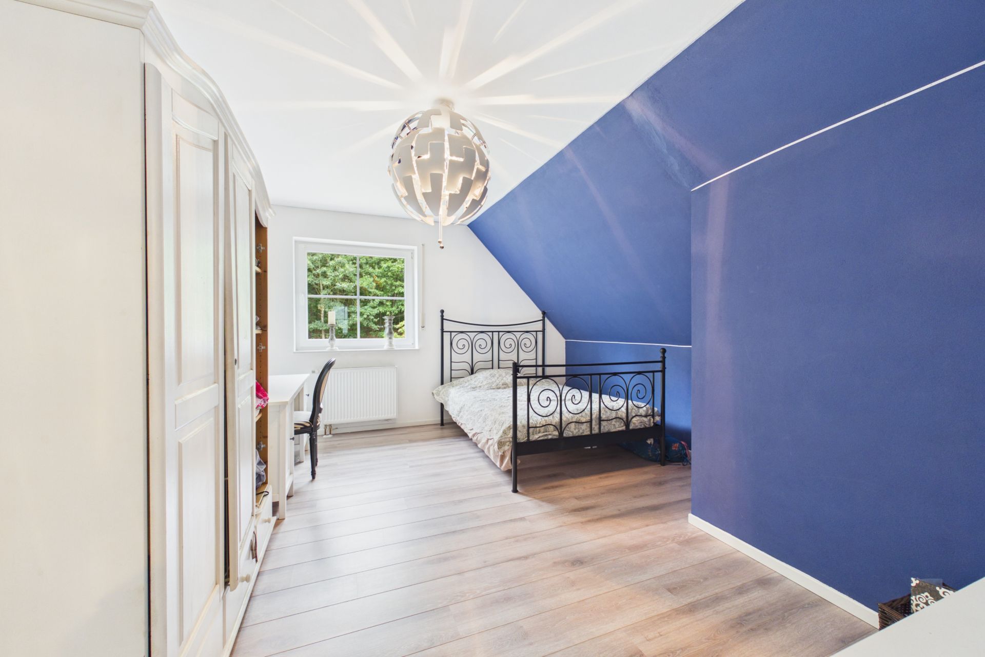 Helles Schlafzimmer mit blau gestrichener Wand, Metallbett, Kleiderschrank und Schreibtisch am Fenster.