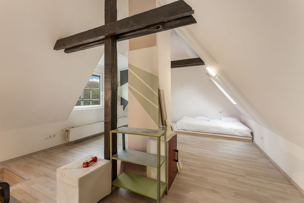 Helles Dachzimmer mit Holzbalken, Regal, Fenster und niedrigem Bett auf dem Boden.