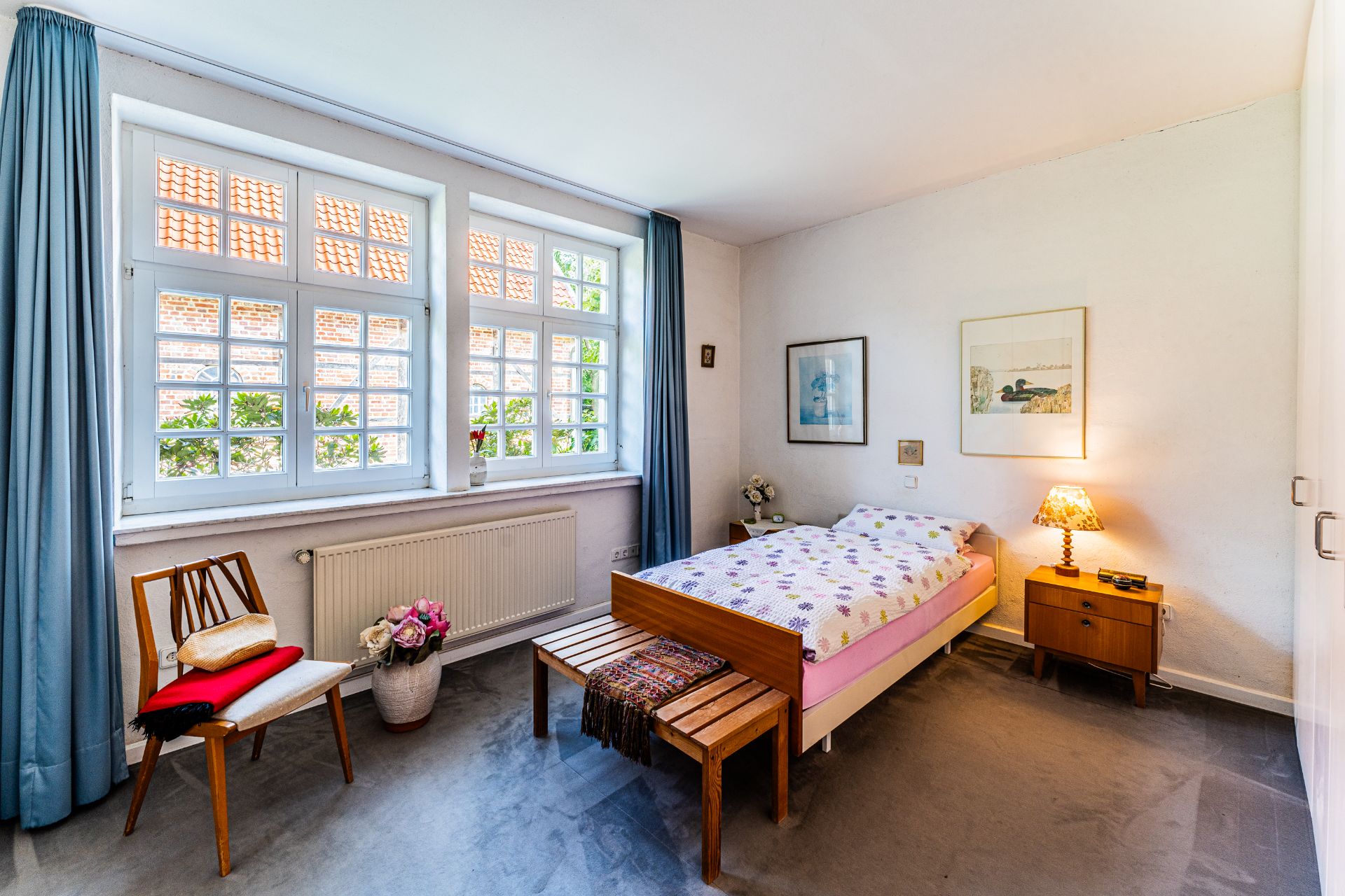 Helles Schlafzimmer mit Einzelbett, Stuhl, Nachttisch, Bank, Blumen und großen Fenstern mit blauen Vorhängen.