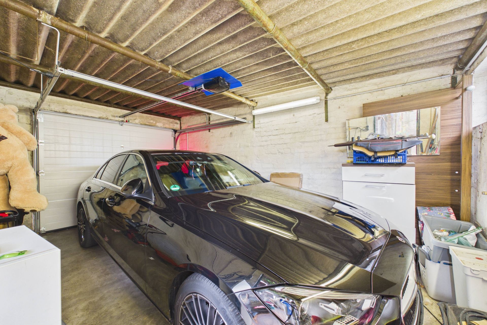 Schwarzes Auto steht in einer Garage mit weißen Wänden und verschiedenen Haushaltsgegenständen.