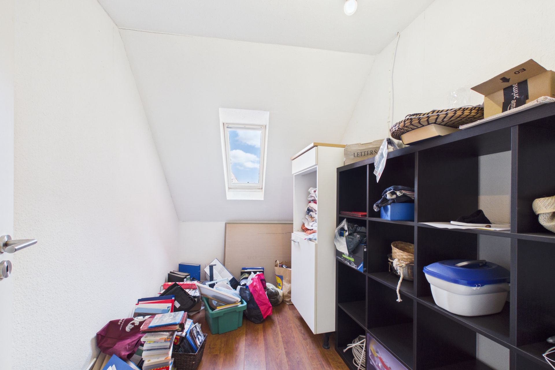 Kleiner Raum mit Regalen, Kartons und verschiedenen Gegenständen, Holzboden, Fenster im Dach.