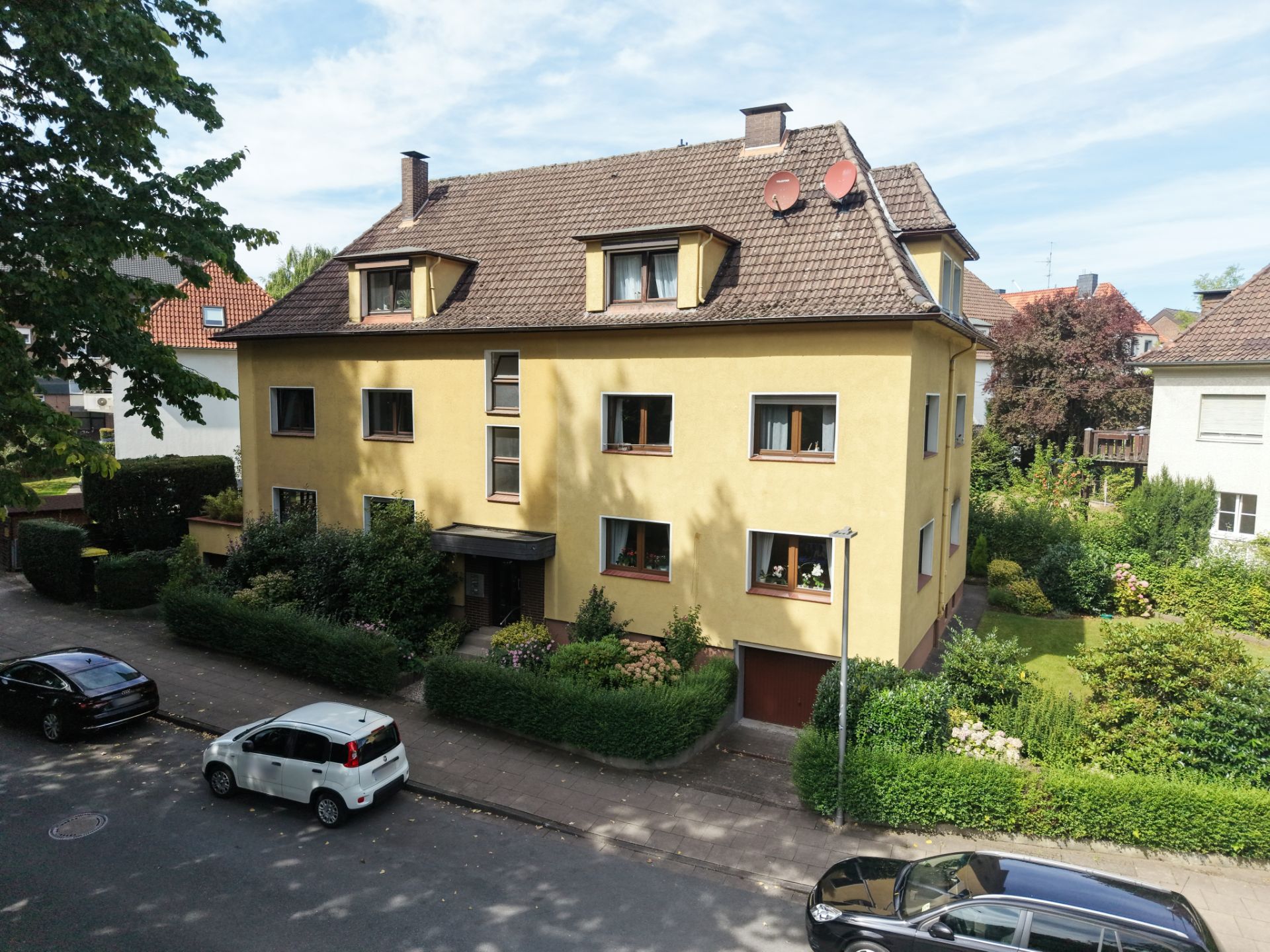 Gelbes Mehrfamilienhaus mit Satteldach, Vorgarten und parkenden Autos an der Straße.
