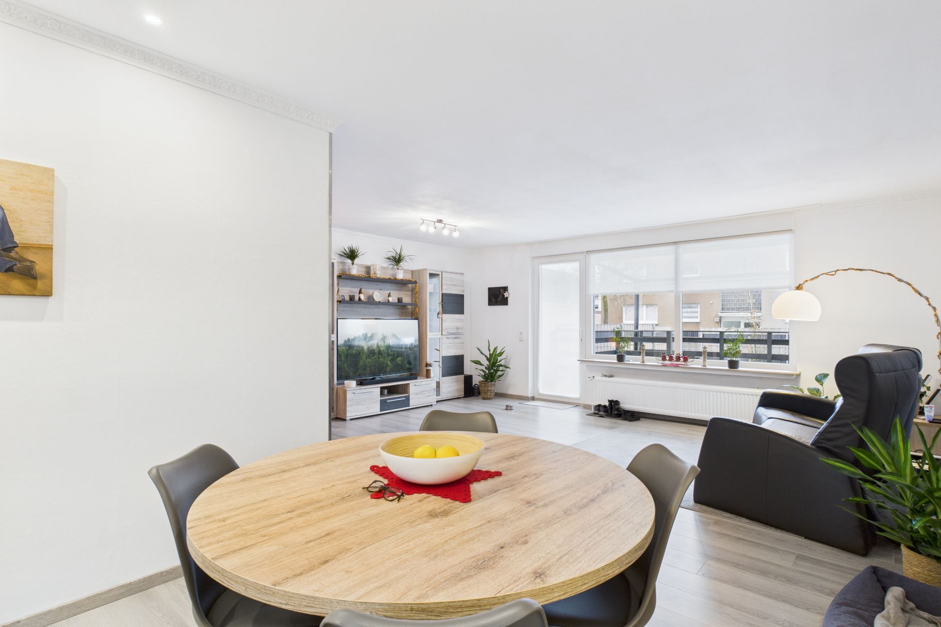Helles Wohnzimmer mit Esstisch, Stühlen, Sofa, Sessel, Fernseher und großen Fenstern.