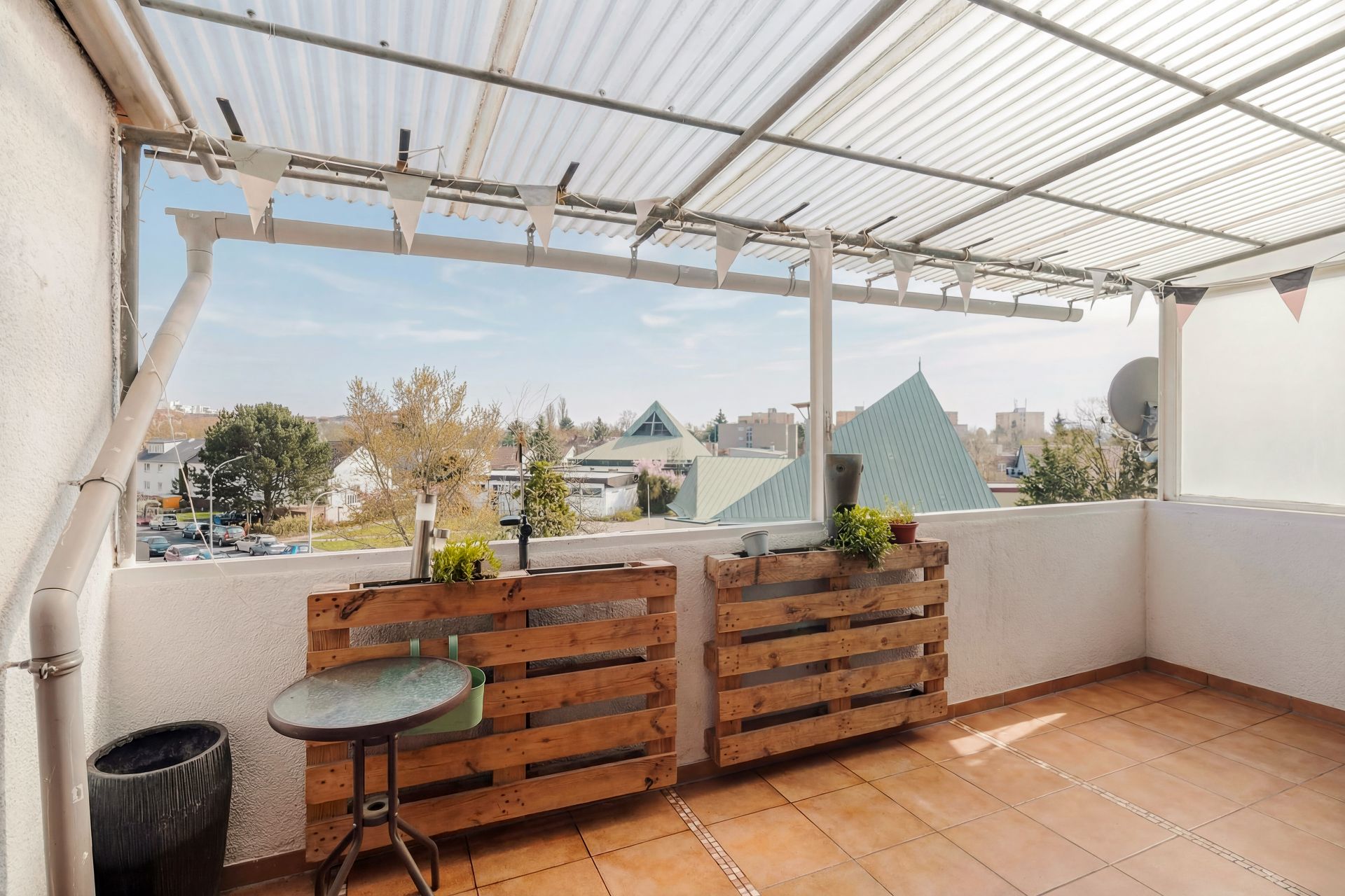 Überdachter Balkon mit Fliesenboden, kleinen Pflanztischen und Blick auf Dächer.
