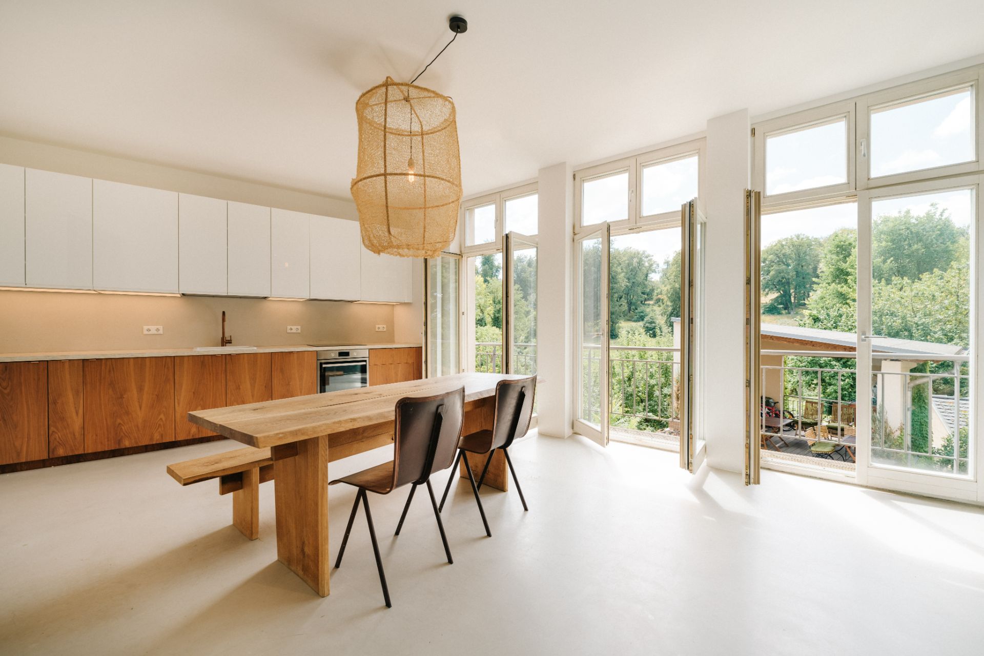 Helle Wohnküche mit Holztisch, Stühlen, großer Fensterfront und Blick ins Grüne.