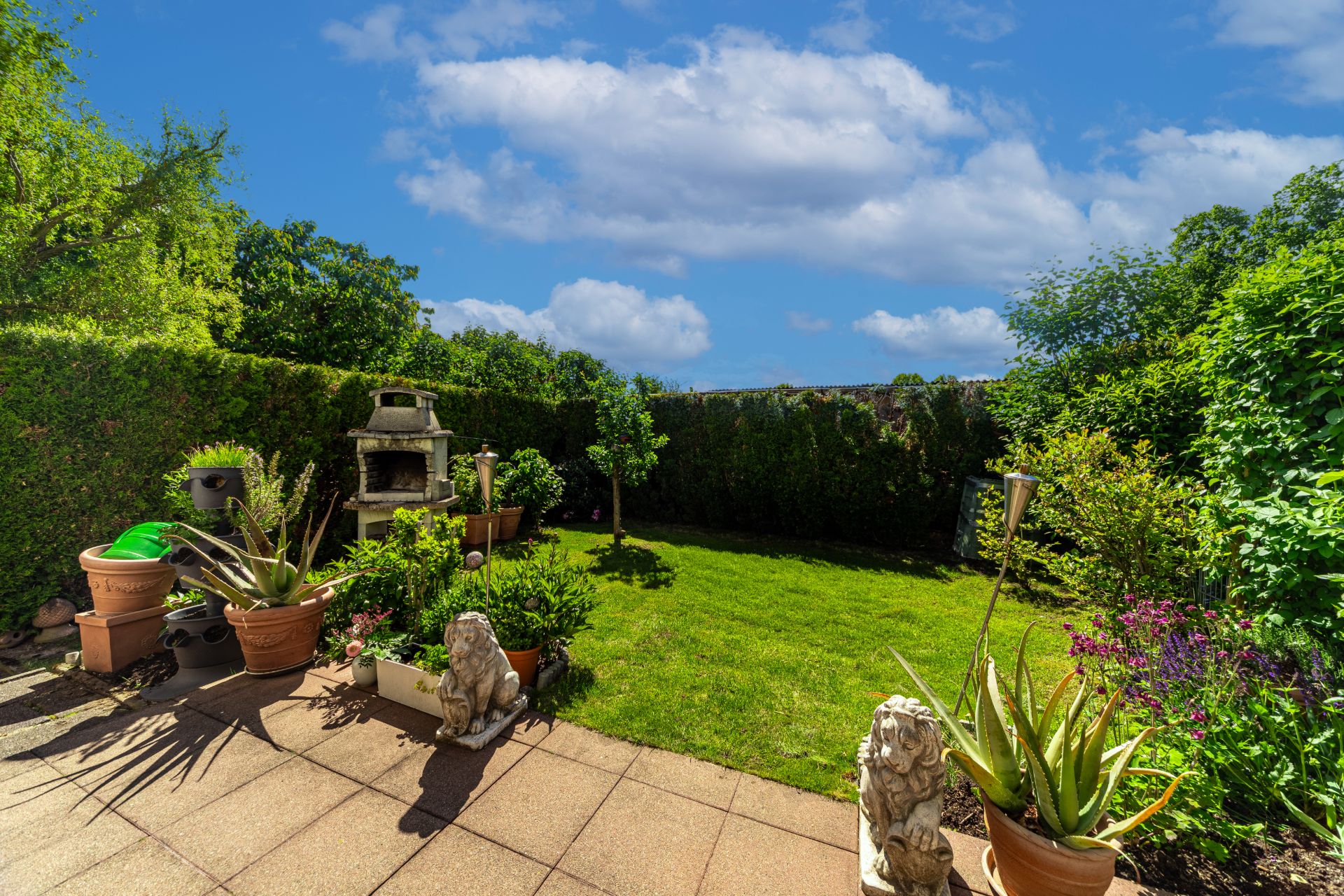 Gepflegter Garten mit Terrasse, Pflanzen in Töpfen und Gartenfigur, umgeben von Hecken.