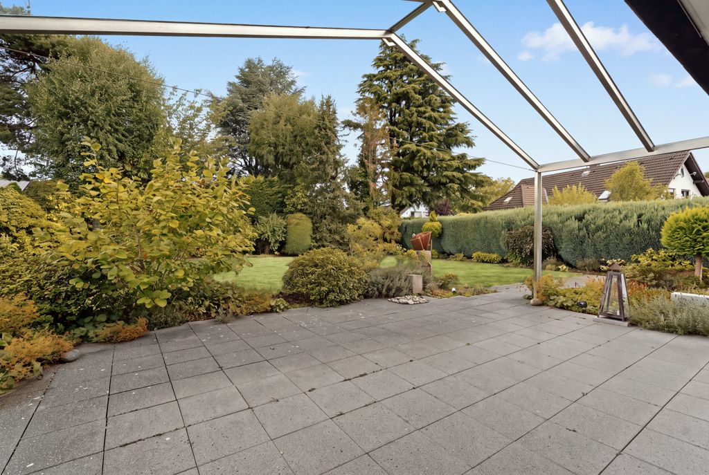Geflieste Terrasse mit Metallrahmen und Blick auf einen grünen Garten mit Sträuchern und Bäumen.