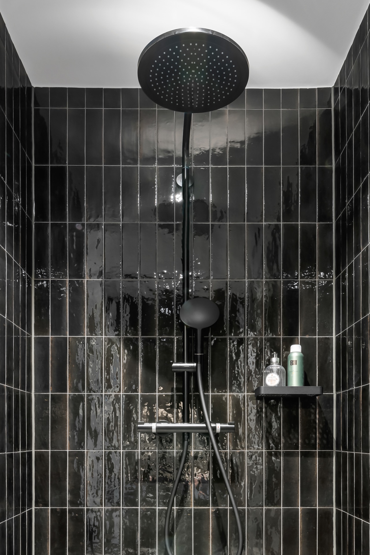 Dusche mit schwarzen Fliesen, Regendusche und kleiner Ablage mit Duschutensilien.