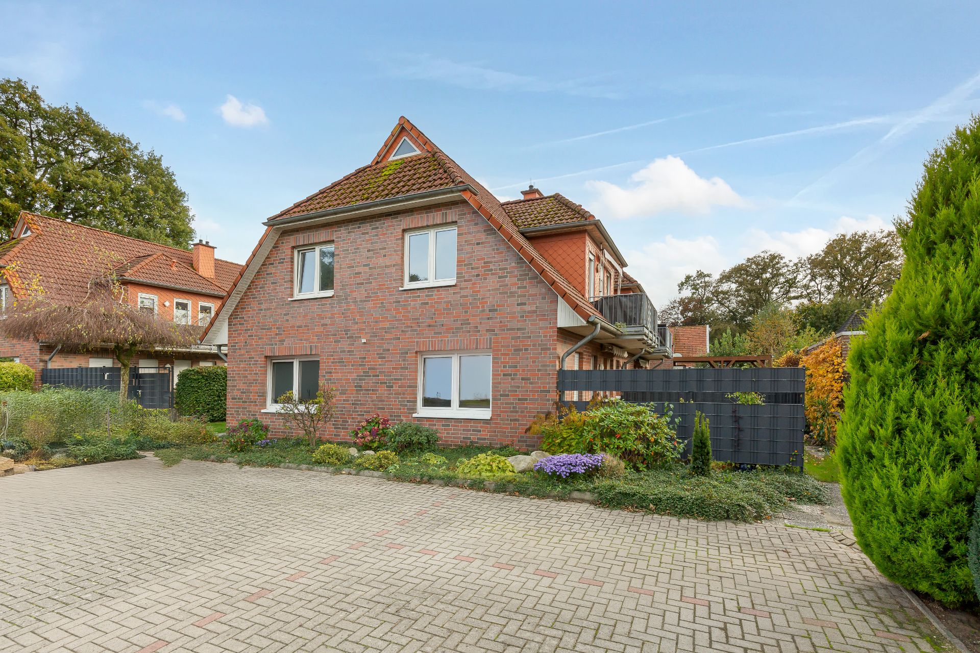 Zweistöckiges Backsteinhaus mit Garten und gepflastertem Hof, umgeben von Bäumen und Sträuchern.