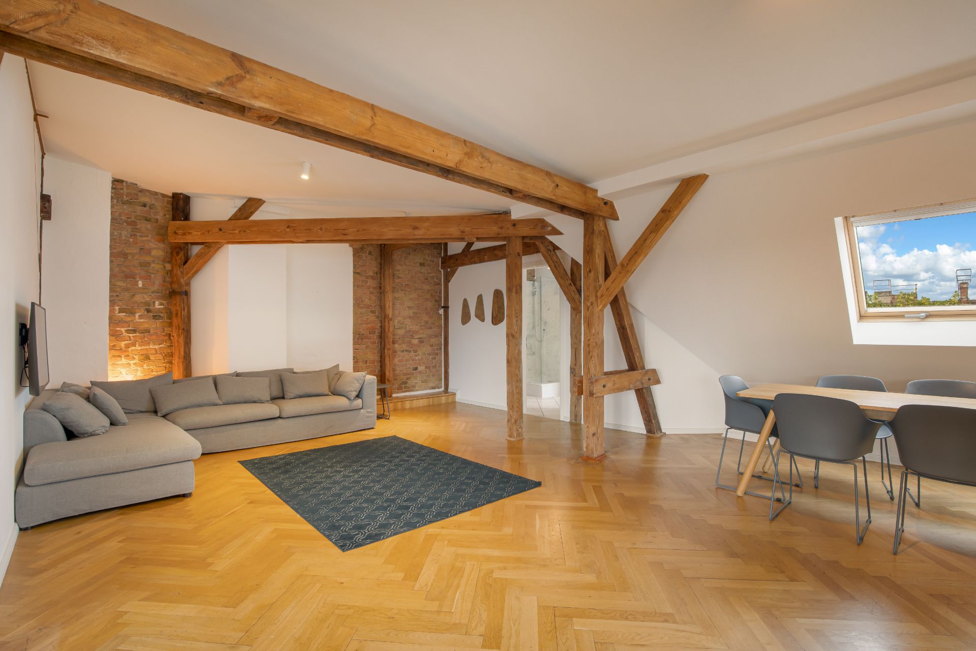 Wohnzimmer mit Holzbalken, grauem Sofa, Teppich, Esstisch und Fenster mit Ausblick.