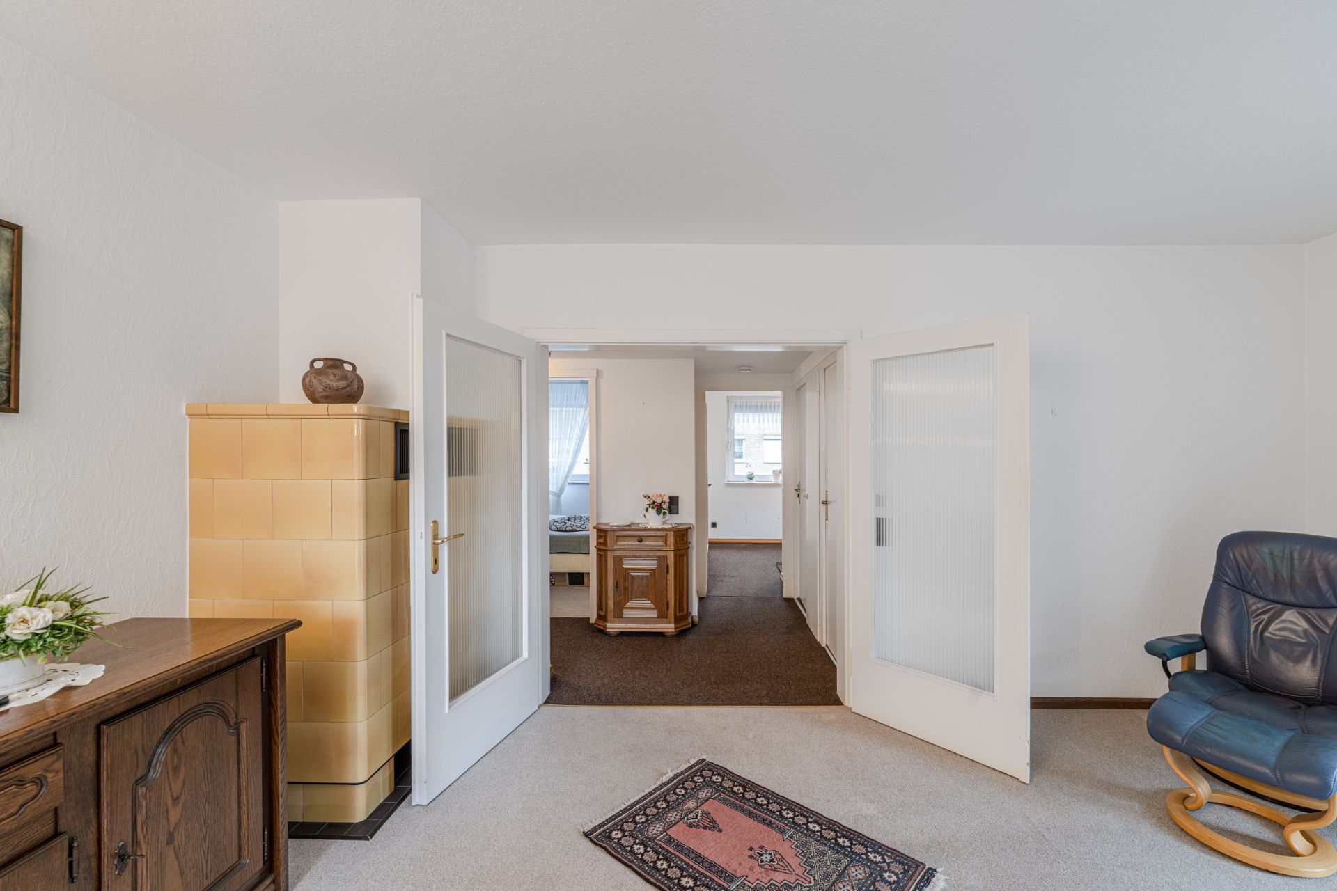 Helles Wohnzimmer mit Teppich, Ofen, Sessel und Blick durch geöffnete Türen auf Flur und Schlafzimmer.