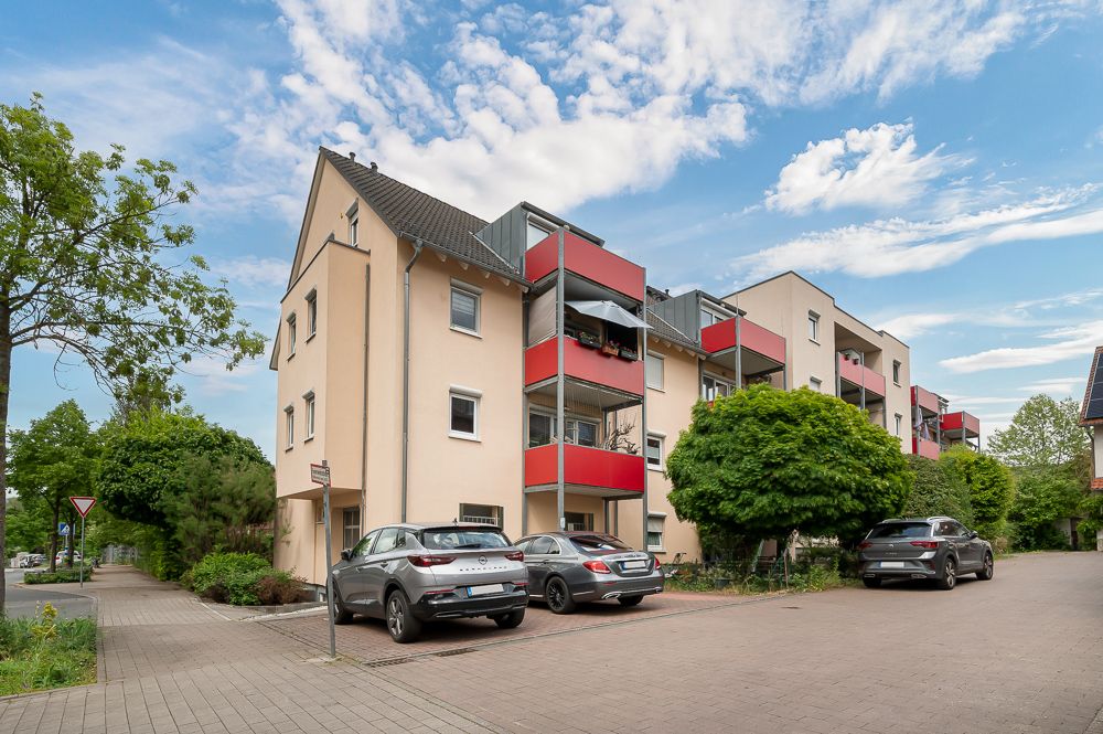 Mehrfamilienhaus mit roten Balkonen, mehreren geparkten Autos und Bäumen im Vordergrund.