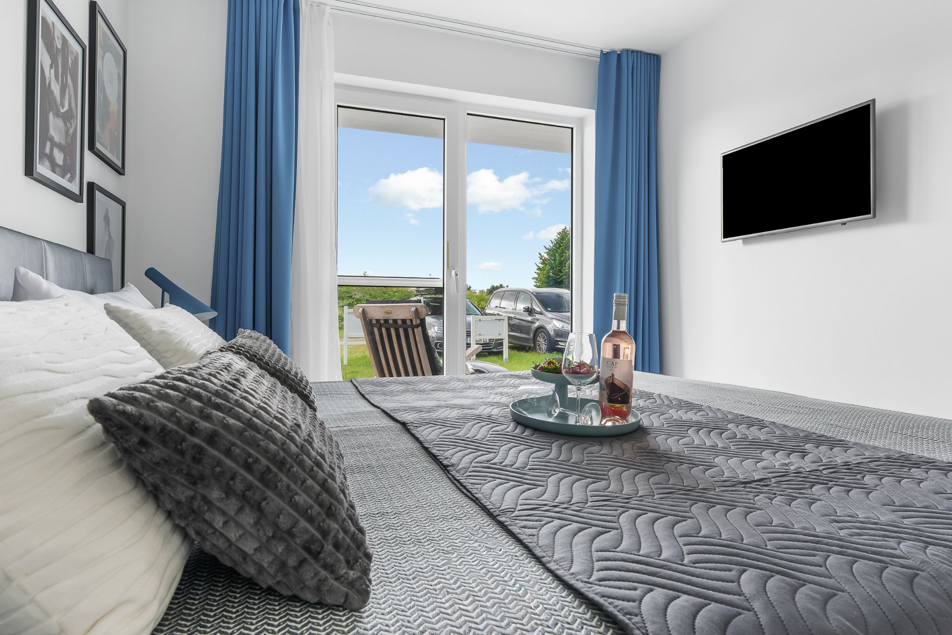 Schlafzimmer mit grauem Bett, Glastür zum Garten, Fernseher an der Wand und blauen Vorhängen.