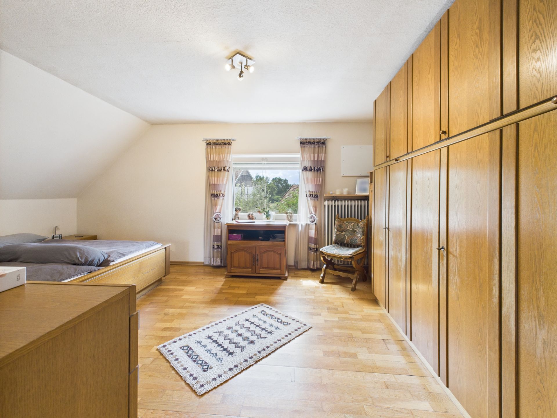 Helles Schlafzimmer mit großem Holzschrank, Bett, Teppich und Fenster mit Vorhängen.