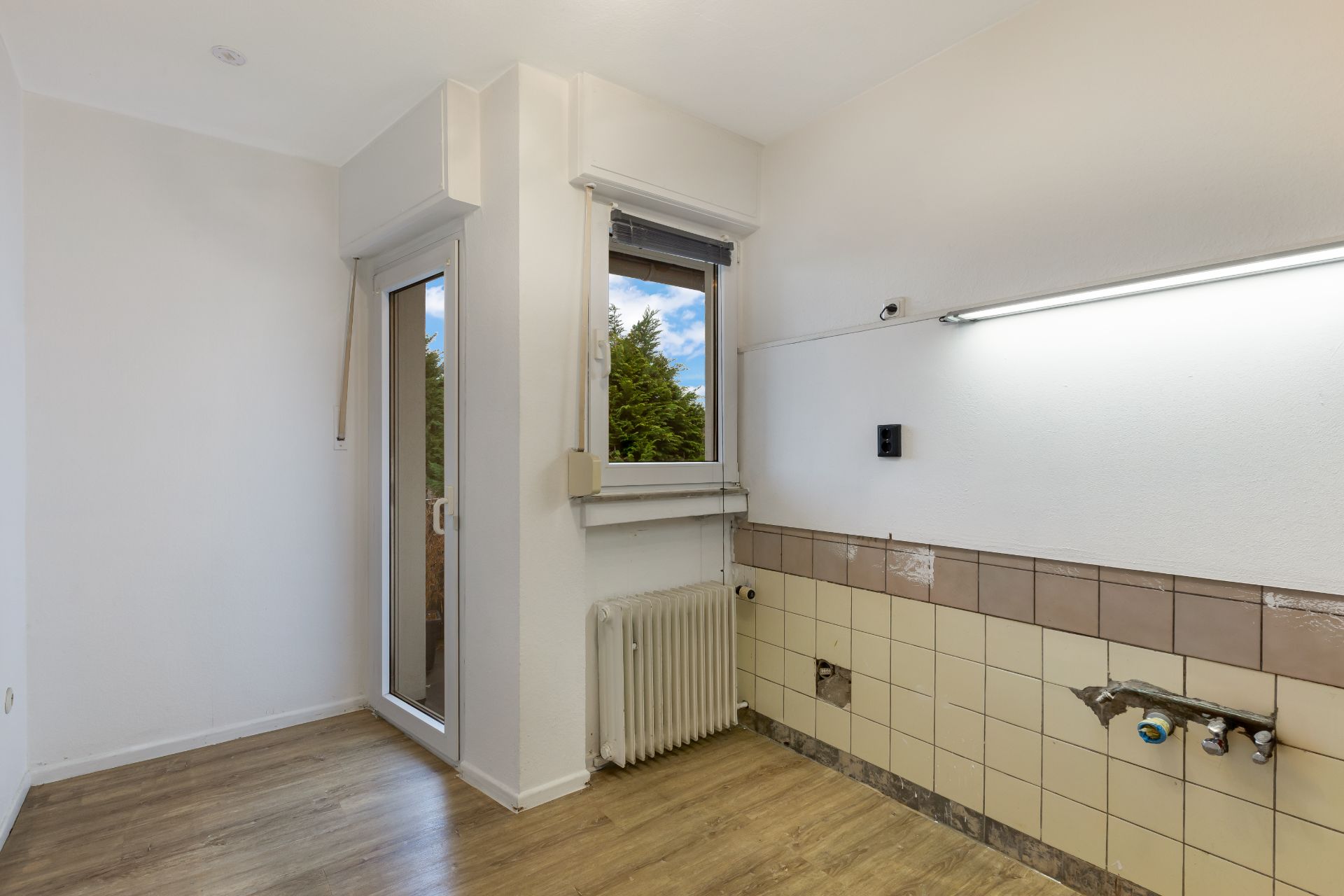 Leerer Raum mit Fenster, Tür zum Balkon, Heizkörper und gefliester Wand mit Wasseranschlüssen.