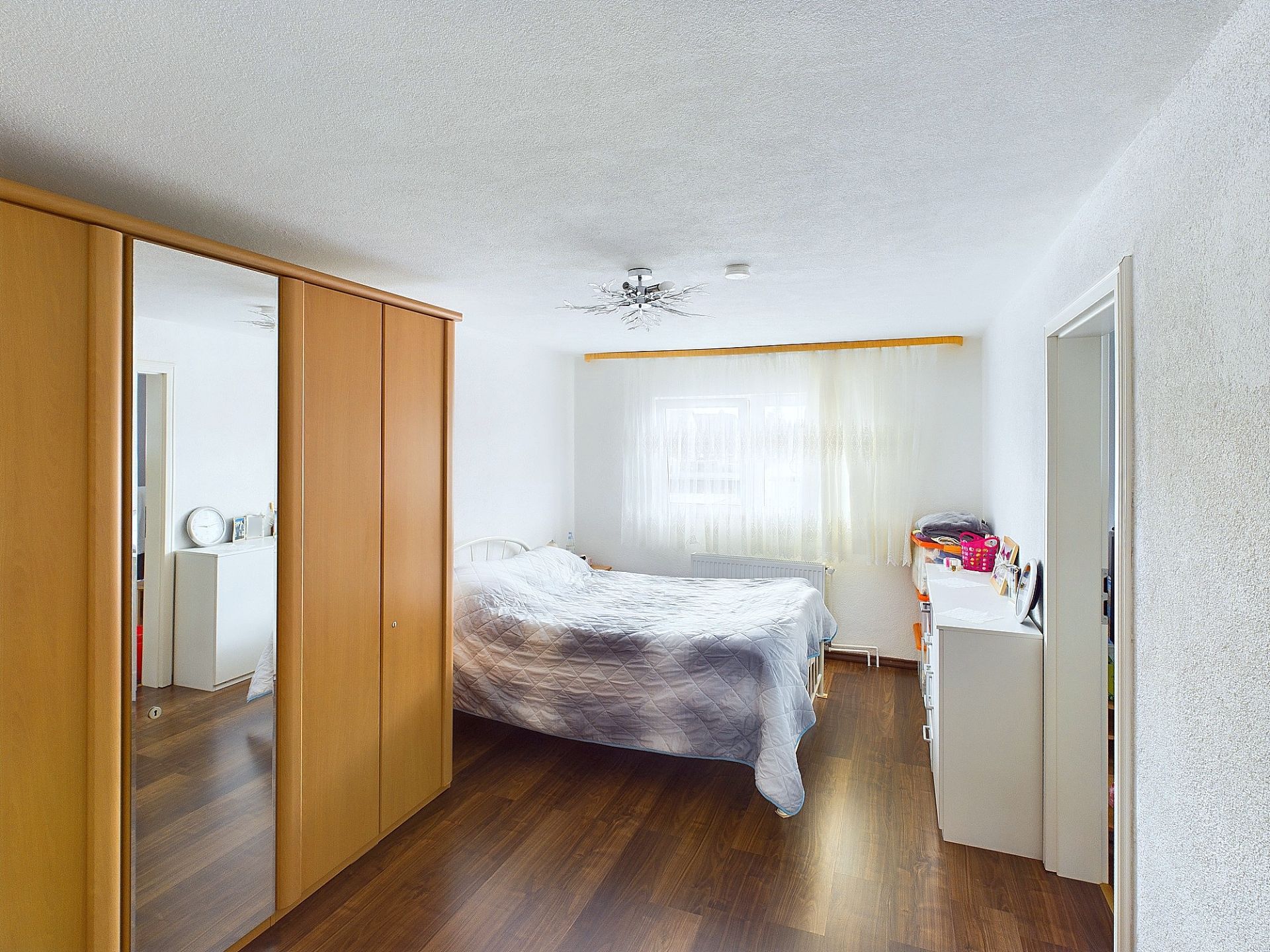 Helles Schlafzimmer mit Doppelbett, Kleiderschrank und Kommode auf dunklem Holzboden.