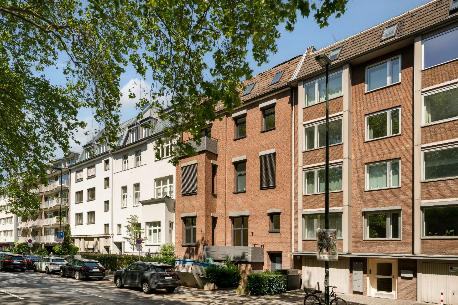 Mehrstöckige Wohnhäuser mit Balkonen und geparkten Autos an einer baumbestandenen Straße.