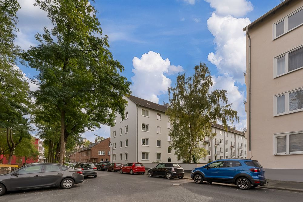 Mehrfamilienhäuser mit geparkten Autos und Bäumen an einer ruhigen Straße.