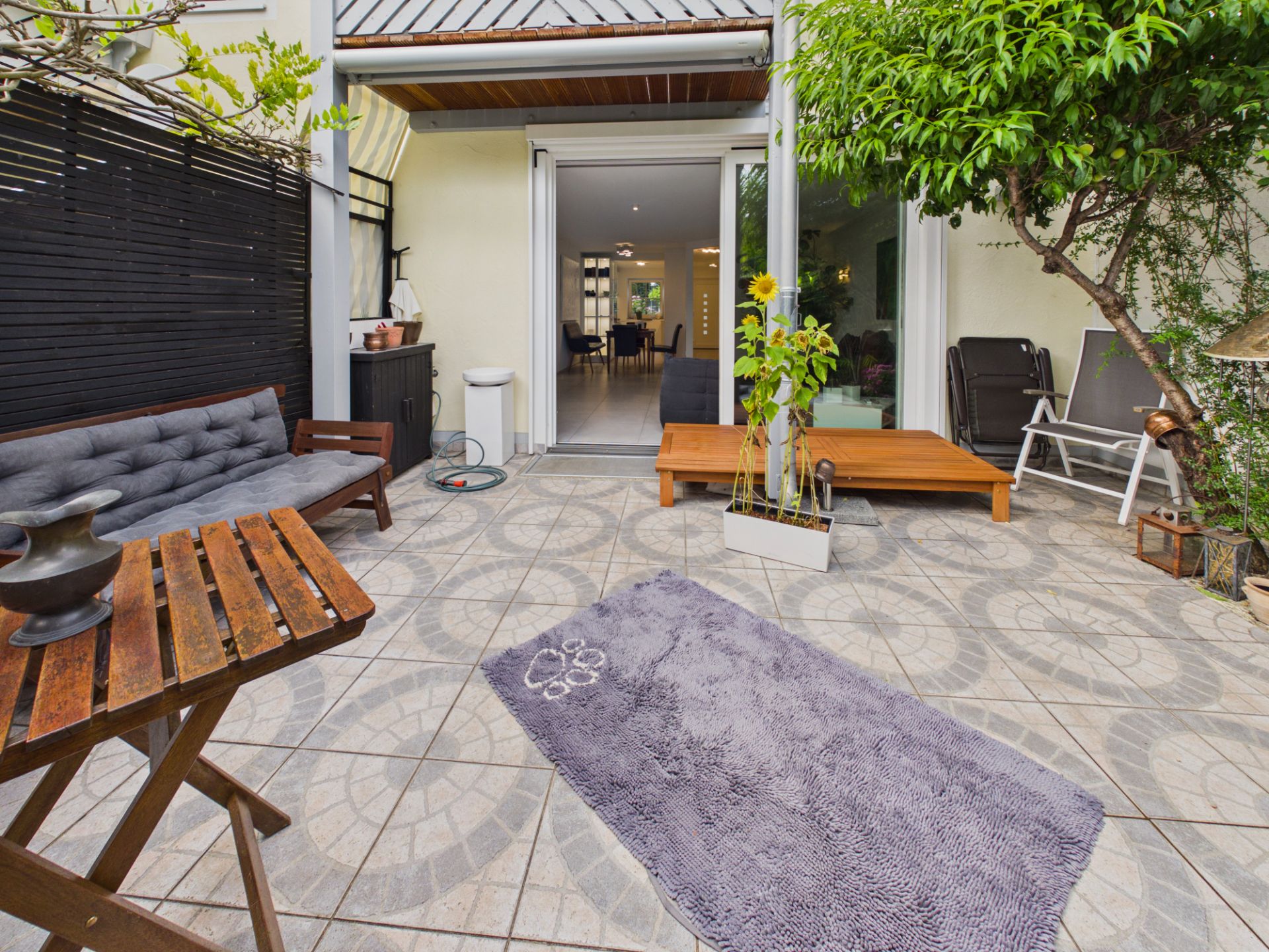 Geflieste Terrasse mit Sitzbank, Tisch, Pflanze im Topf und Zugang zum Wohnzimmer.