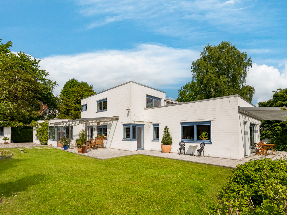 Modernes weißes Wohnhaus mit flachem Dach, großen Fenstern und Terrasse im Garten.