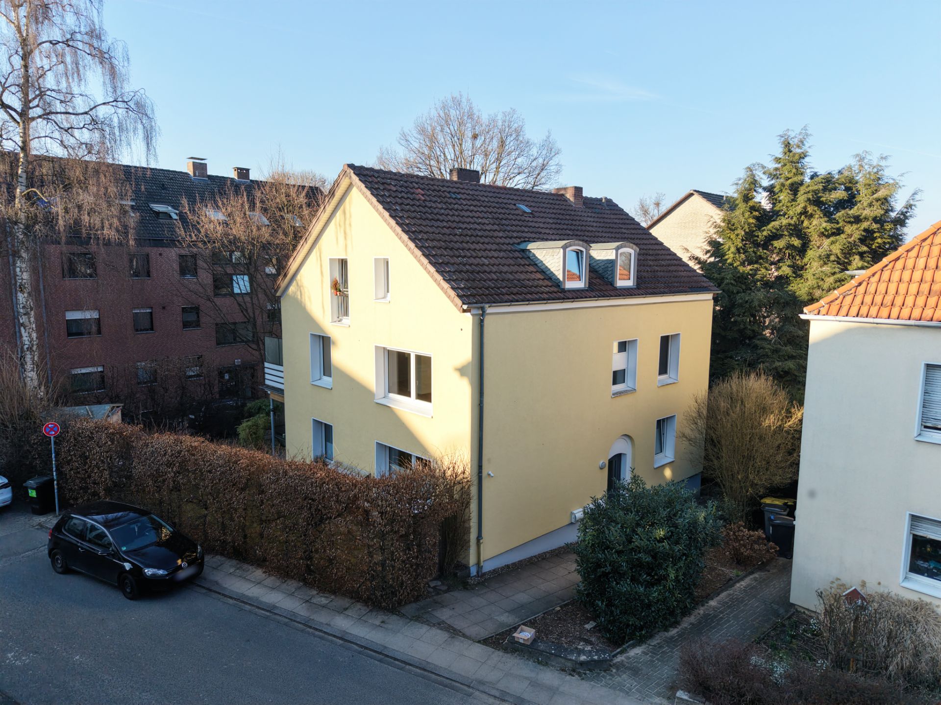 Gelbes Wohnhaus mit rotem Dach an einer Straßenecke, umgeben von Hecken und Nachbargebäuden.