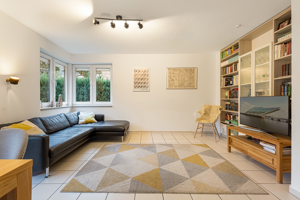 Helles Wohnzimmer mit Ecksofa, Teppich, Bücherregal und Fernseher auf Holzschrank.