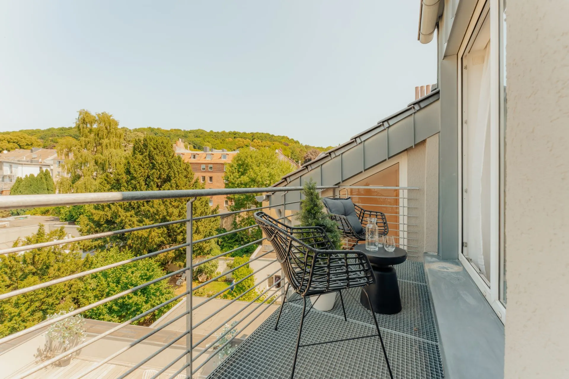 Balkon mit zwei Stühlen, kleinem Tisch und Pflanzen, Blick auf Bäume und Gebäude.