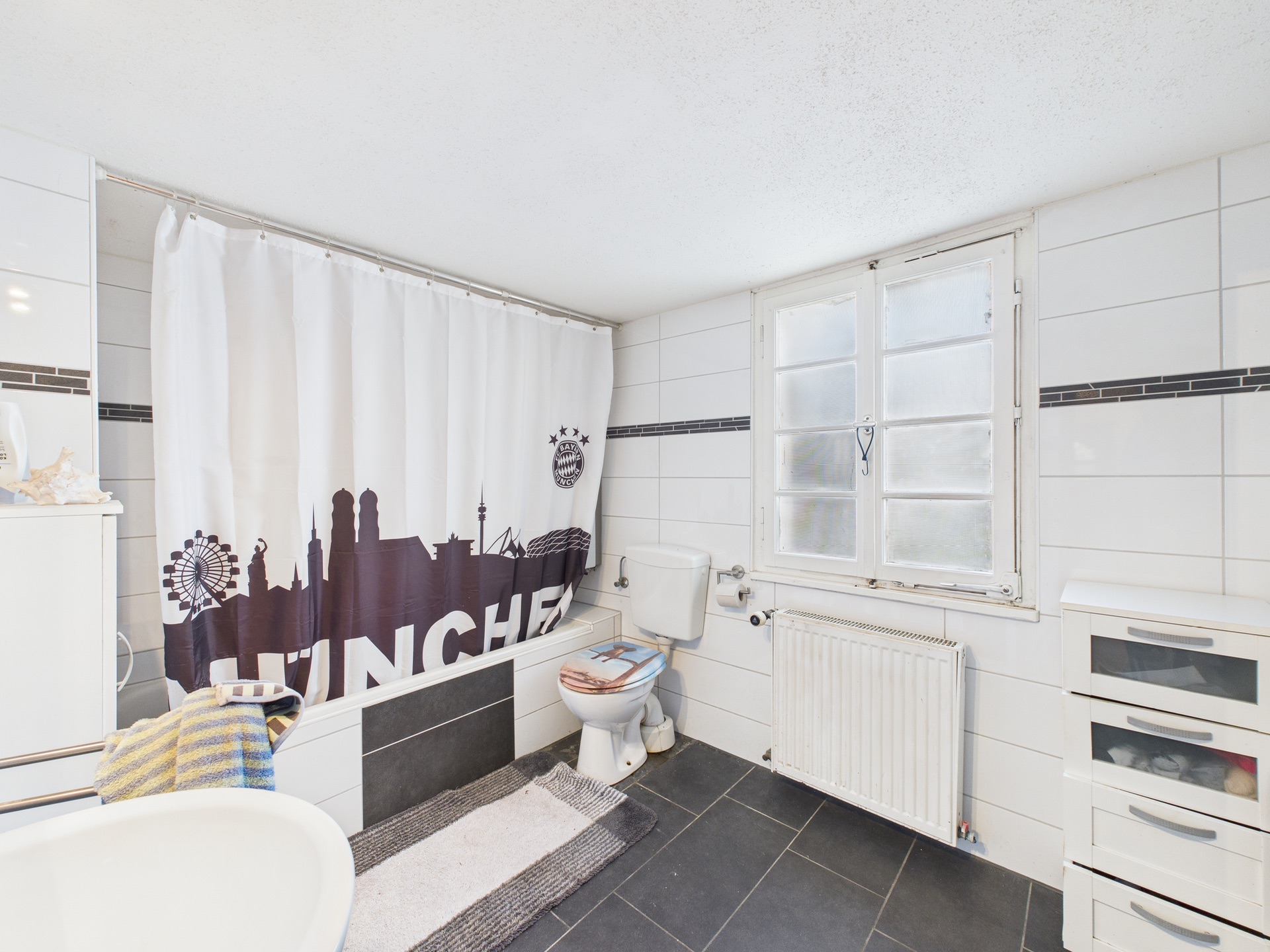 Helles Badezimmer mit Fenster, Badewanne, Duschvorhang, Toilette und weißen Fliesen.