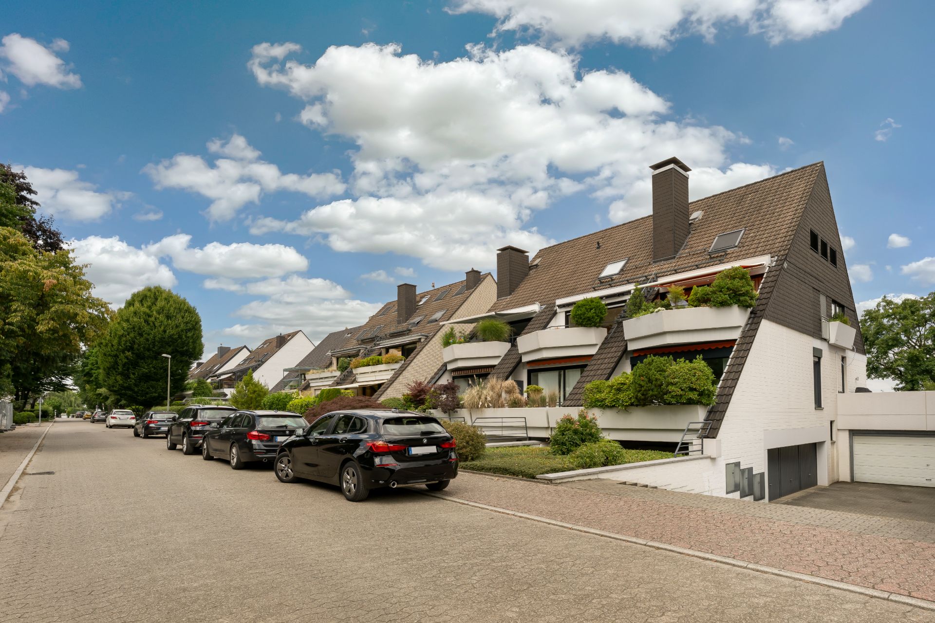 Mehrfamilienhaus mit schrägen Dächern und Balkonen, davor geparkte Autos an einer ruhigen Straße.