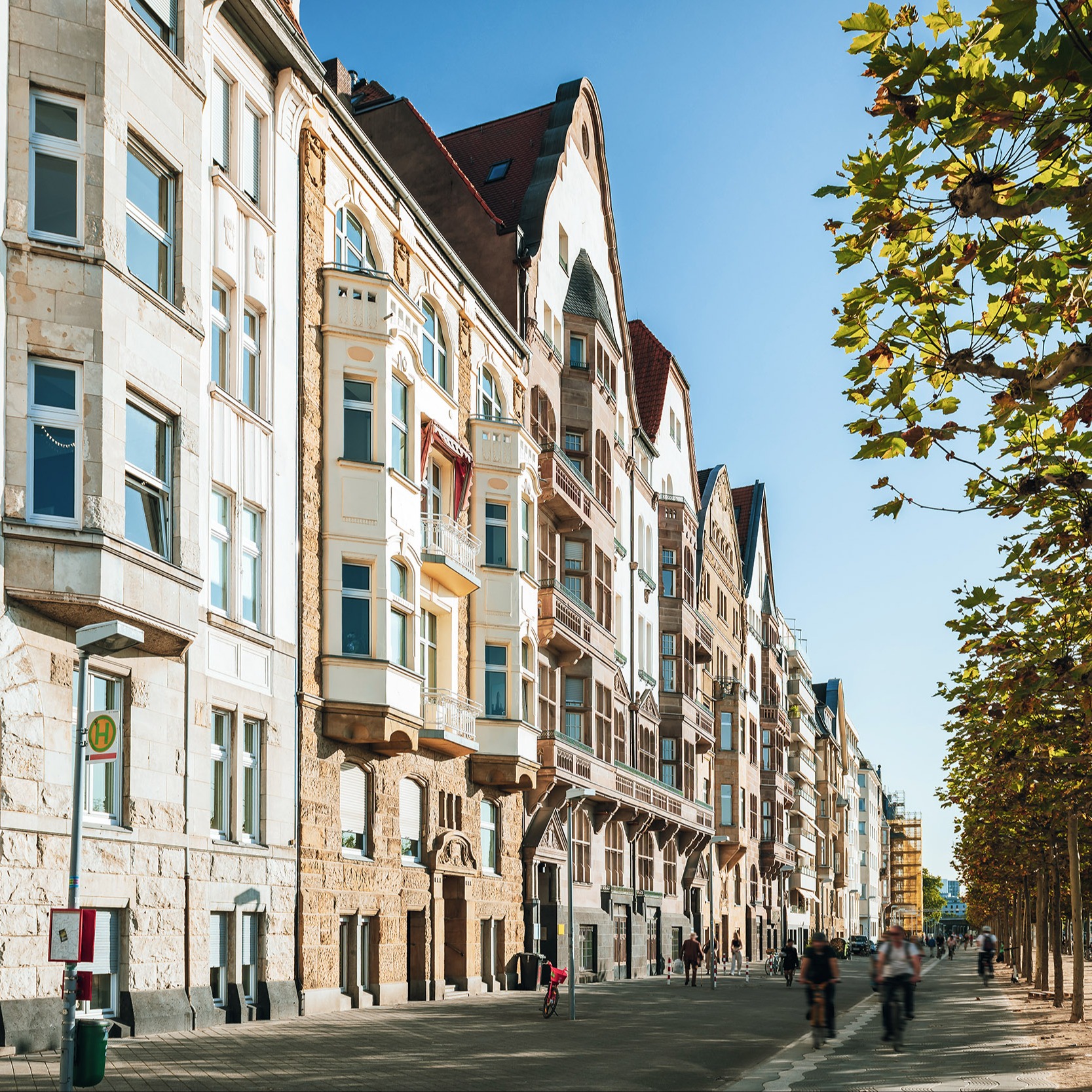Mehrstöckige Wohnhäuser mit dekorativen Fassaden an einer breiten Straße mit Bäumen und Fußgängern.