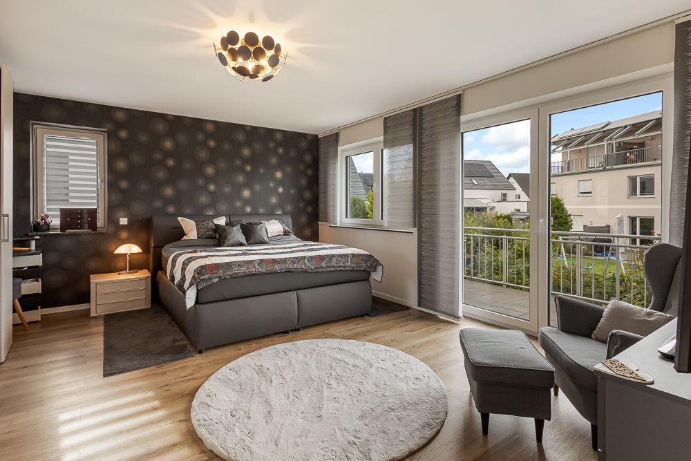 Modernes Schlafzimmer mit Doppelbett, Sessel, rundem Teppich und großen Fenstern zu einem Balkon.