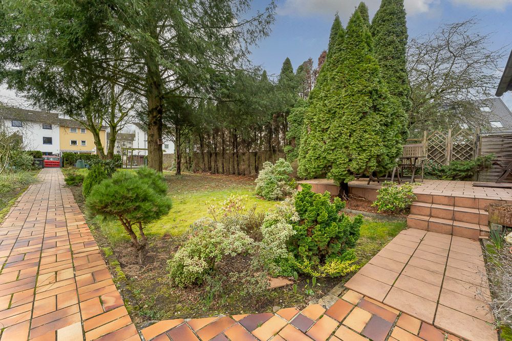 Gepflasterter Weg und Terrasse im Garten mit Bäumen, Sträuchern und Sitzgelegenheit.