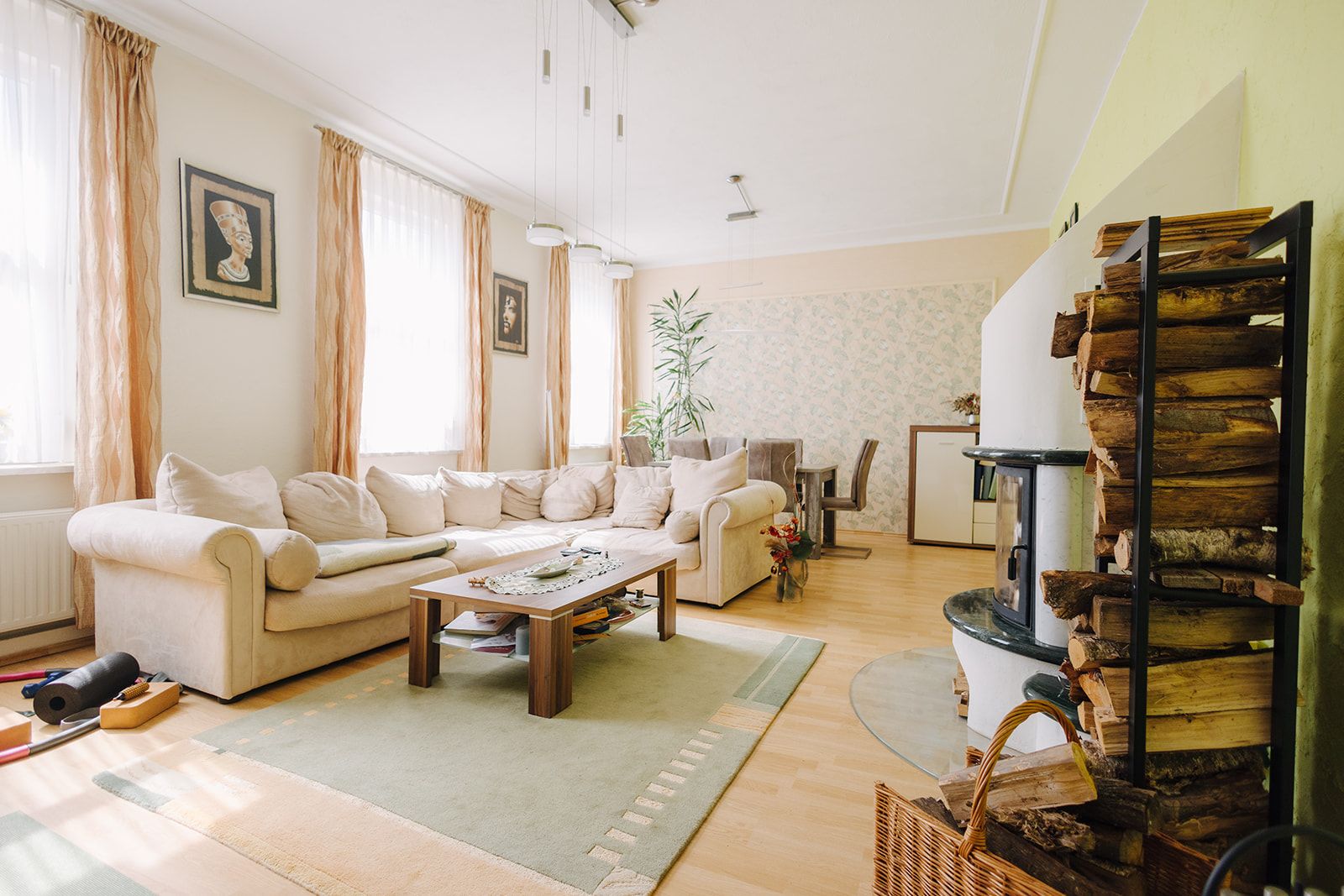 Helles Wohnzimmer mit beiger Eckcouch, Couchtisch, Teppich, Holzofen und gestapeltem Feuerholz.