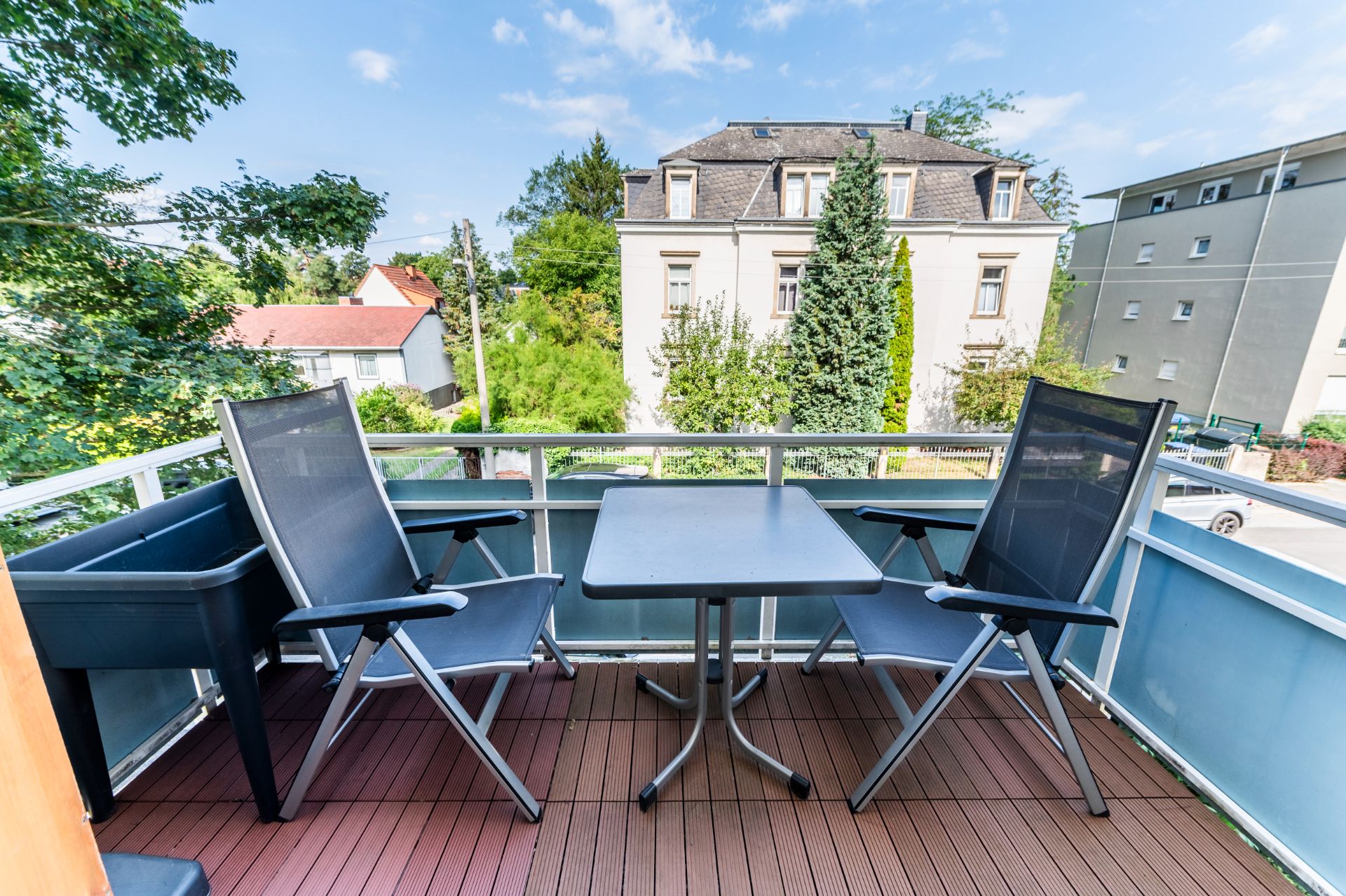 Balkon mit zwei Stühlen, einem Tisch und Blumenkasten, Blick auf Wohnhäuser und Bäume.
