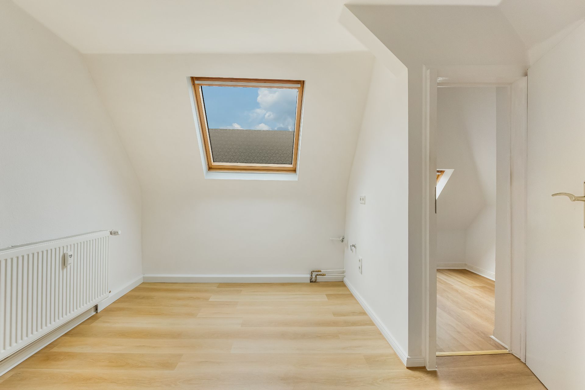 Heller Raum mit Dachfenster, Laminatboden und Radiator, Tür führt in weiteren ähnlichen Raum.
