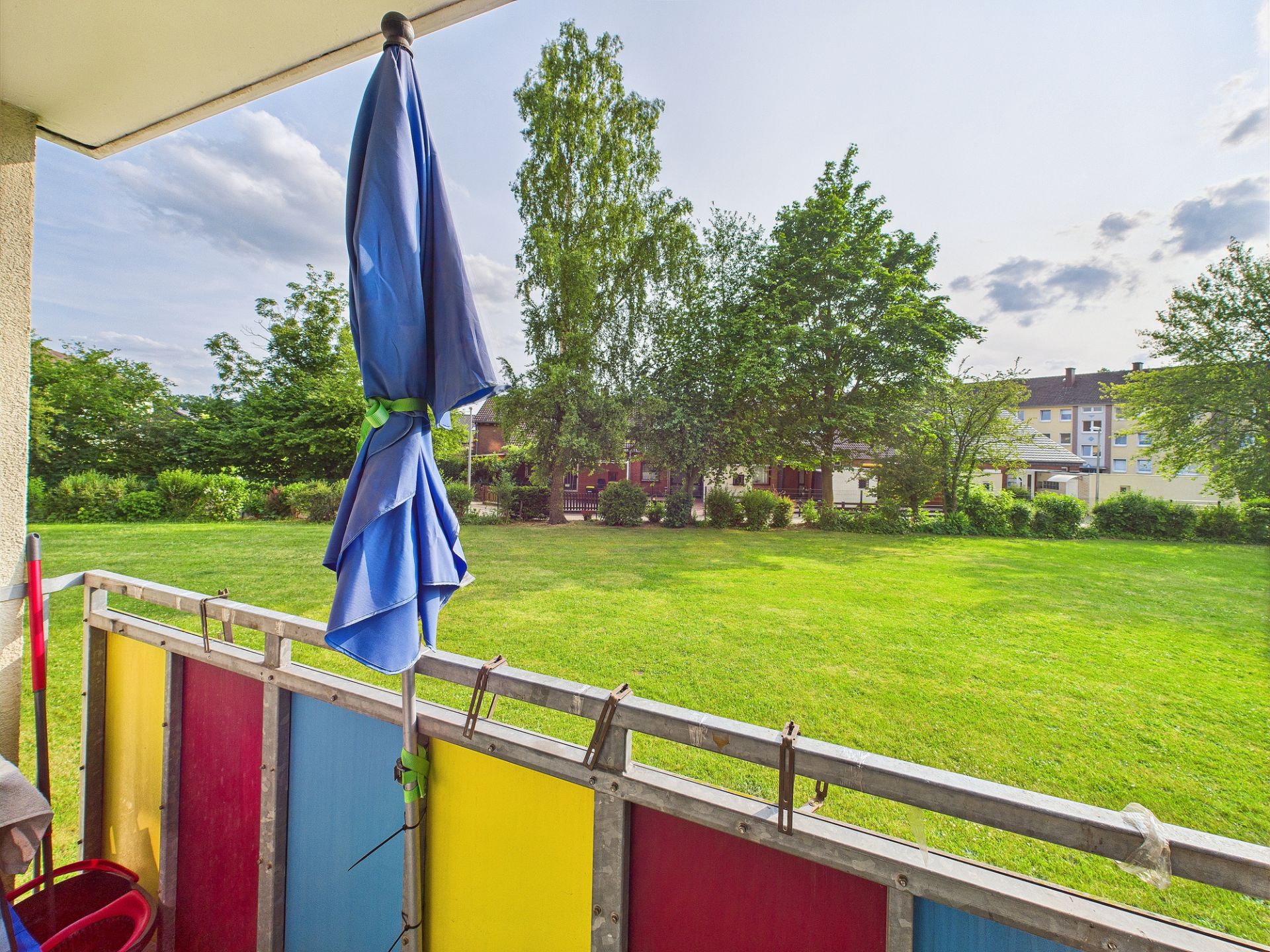 Blick vom Balkon mit buntem Geländer auf eine große grüne Wiese und Bäume.