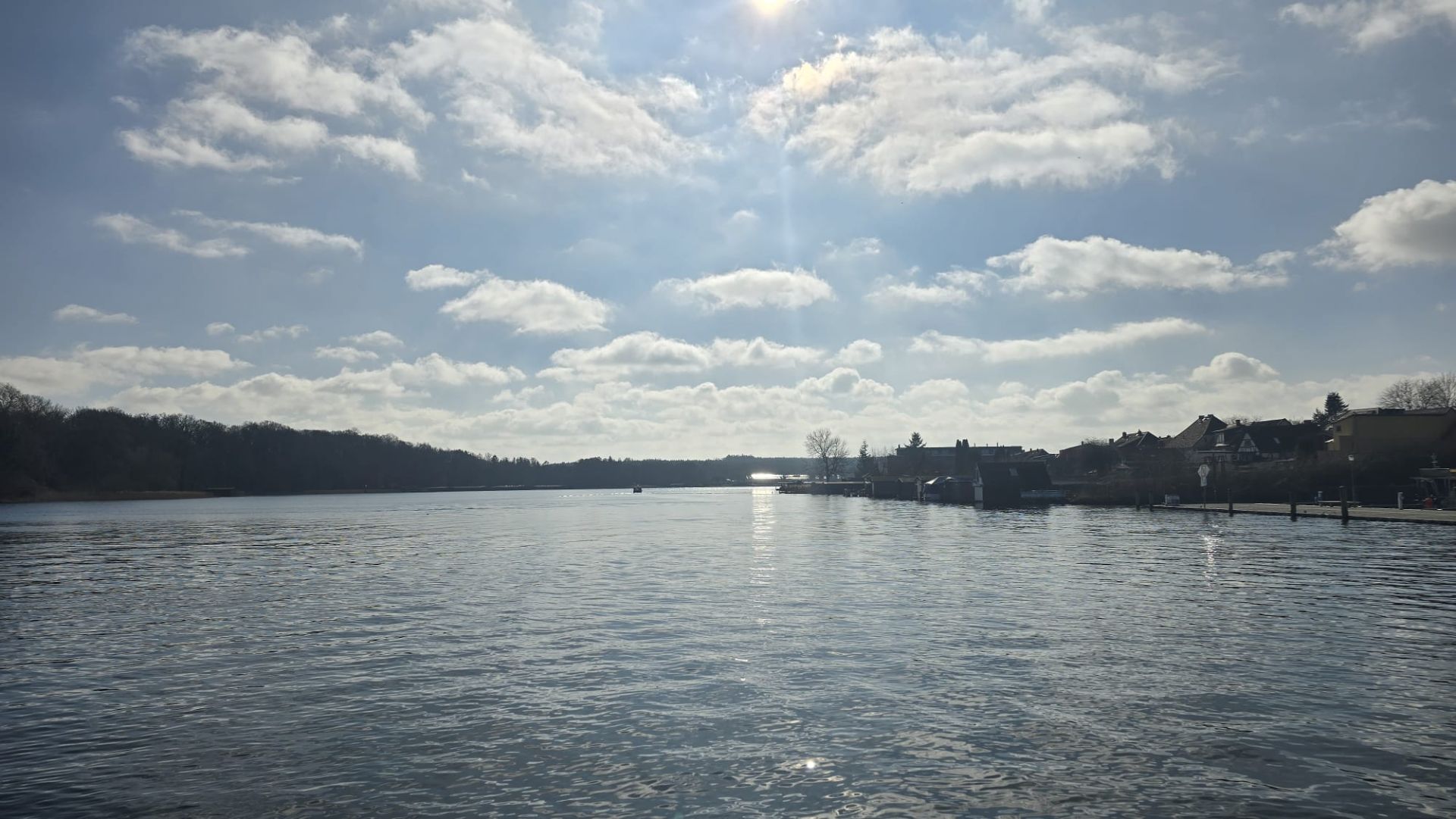 Blick auf einen ruhigen See mit Häusern am rechten Ufer unter bewölktem Himmel.