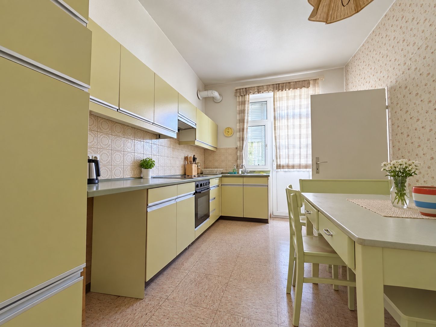 Helle Küche mit gelben Schränken, Esstisch mit Stühlen und Fenster mit Vorhang.