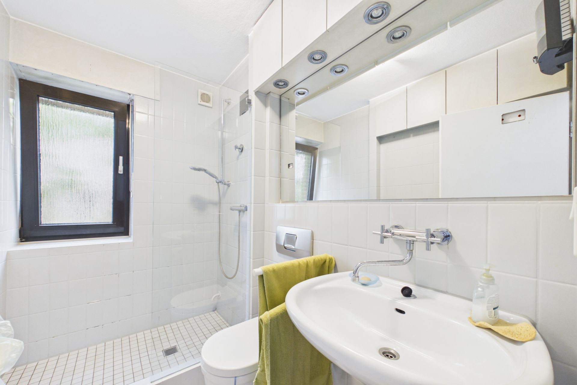 Helles Badezimmer mit Fenster, Dusche, Waschbecken, Spiegel und Handtuch.