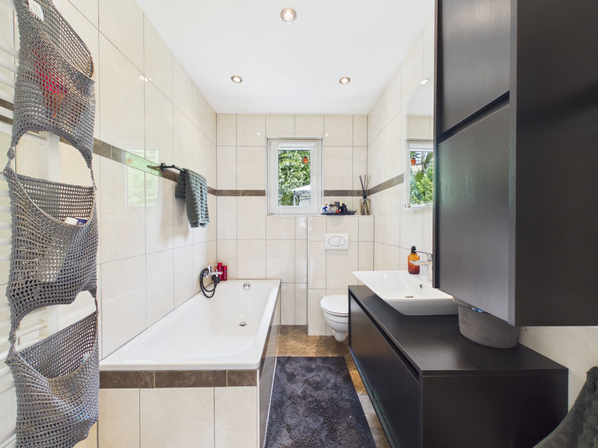 Modernes Badezimmer mit Badewanne, Waschbecken, WC und zwei Fenstern.